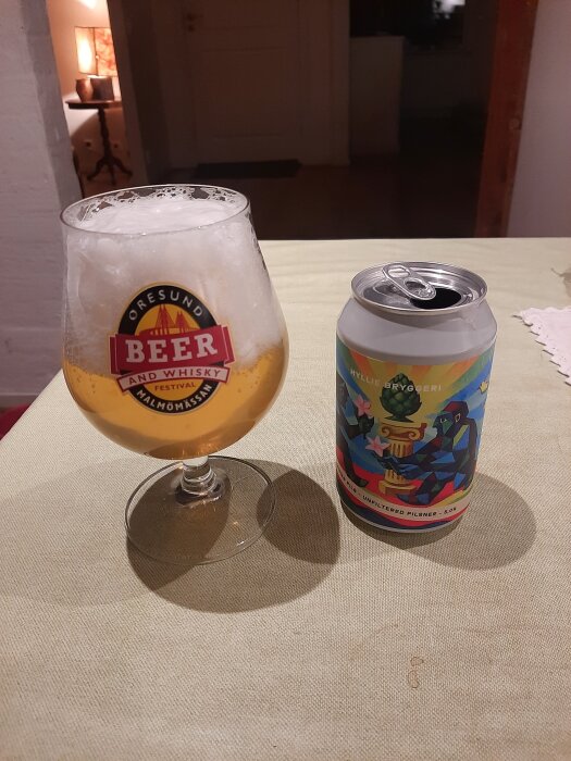 Ett glas öl med skum intill en färgglad ölburk på ett bord.