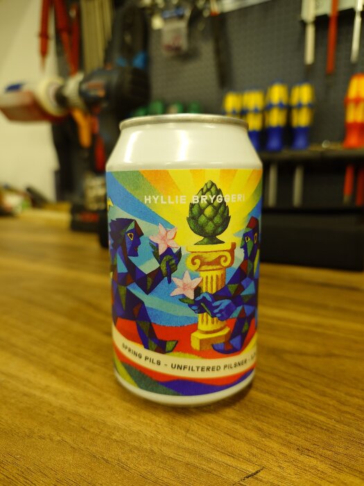 Ölburk med etikett från Hyllie Bryggeri, motiv med geometriska figurer och text "Spring Pils - Unfiltered Pilsner" på ett träbord.