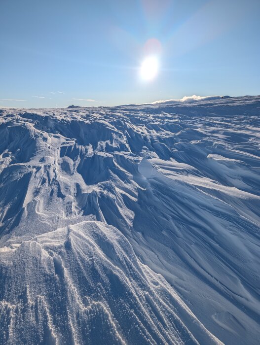 Snötäckt landskap med solen högt på himlen, vilket skapar långa skuggor över de snötäckta åsarna.