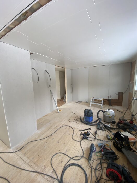 Renovering pågår i ett rum med verktyg och dammsugare på ett trägolv, samt nydragen el i taket och väggar klara för ny beklädnad.