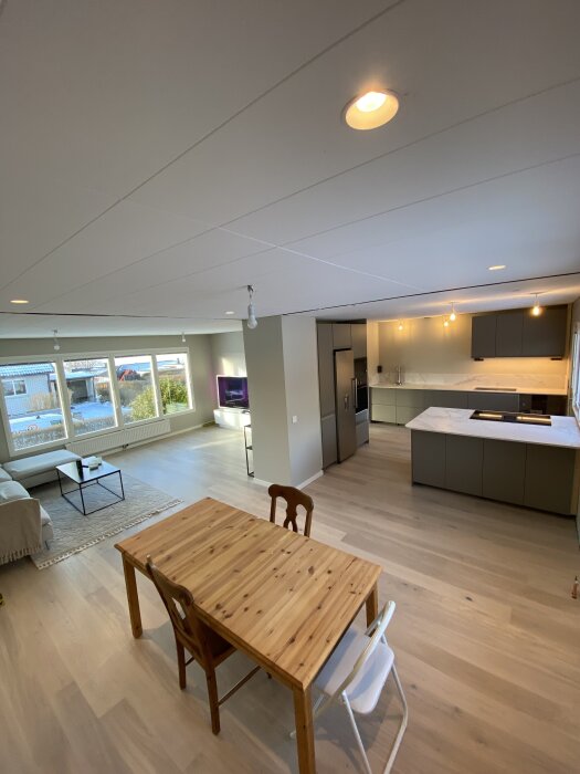 Öppet vardagsrumskoncept med matbord och stolar i trä, modernt kök med grå skåp, vit köksö och soffhörna med stora fönster i ett Myresjöhus.