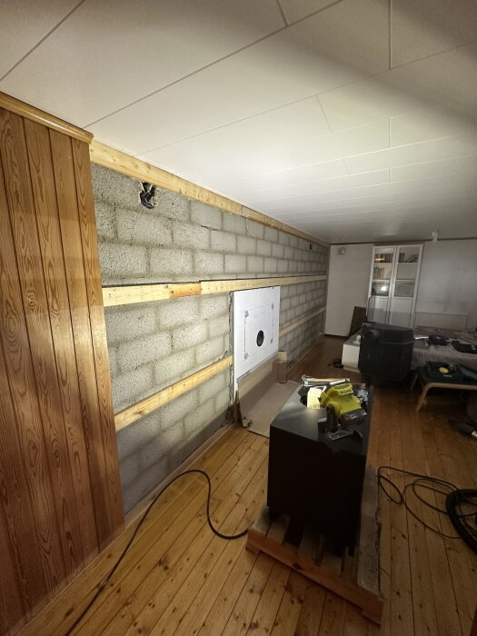 Renovering av gillestuga med blottad stenvägg och ny infälld kamin. Panel och tapet har tagits bort, och vedförvaring byggts in i väggen.