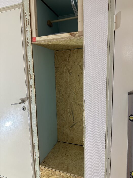 Öppen garderob under renovering med gipsvägg och OSB-bjälkar, vit dörr bredvid.