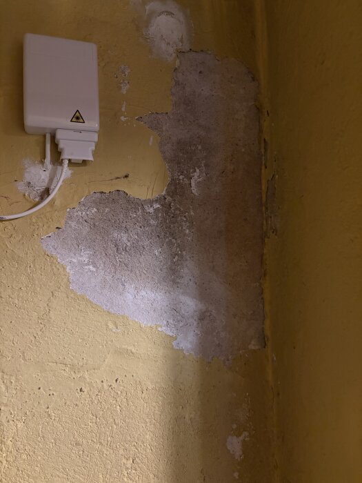 Gul vägg där puts och färg har släppt, avslöjar underliggande yta. Vit elektrisk låda monterad ovanför skadad del av väggen.