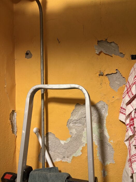 Skadad vägg med flagnande gul färg och exponerad puts. Stege och en vit stav står framför väggen.