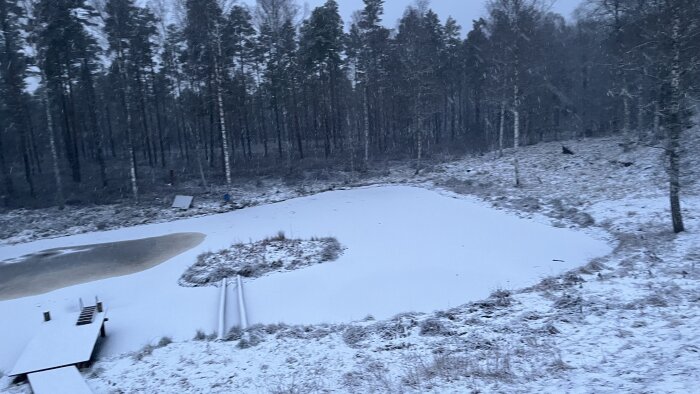 Snötäckt damm med brygga omgiven av skog. Vinterscen där isen har lagt sig över vattnet, visar stillhet och naturens vila.