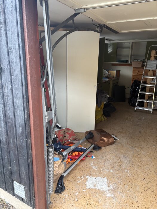 Öppen garageport med viparmsystem, omgiven av oordning med verktyg och väskor, i ett garage med hyllor och stege i bakgrunden.