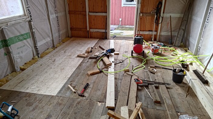 Renoveringsprojekt med träplankor, verktyg och isolering i ett halvt färdigt rum. En sladd och en såg syns på golvet.