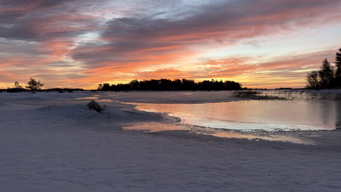 Snötäckt landskap vid en frusen sjö, soluppgång med dramatiska färger i himlen och reflektioner i isen.