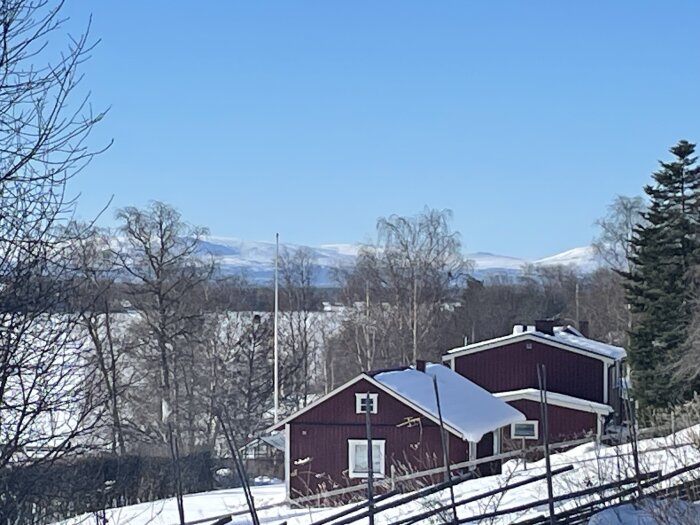 Röda stugor med snötäckta tak i en vintrig landsbygd, omgivna av träd och snöklädda berg i bakgrunden under en klarblå himmel.