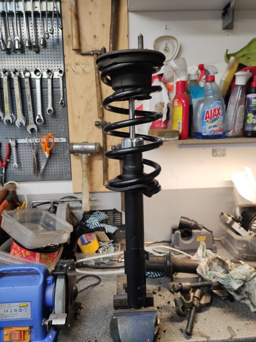 Bilstötdämpare fastspänd i ett skruvstäd på en arbetsbänk i en verkstad, omgiven av verktyg och rengöringsprodukter.