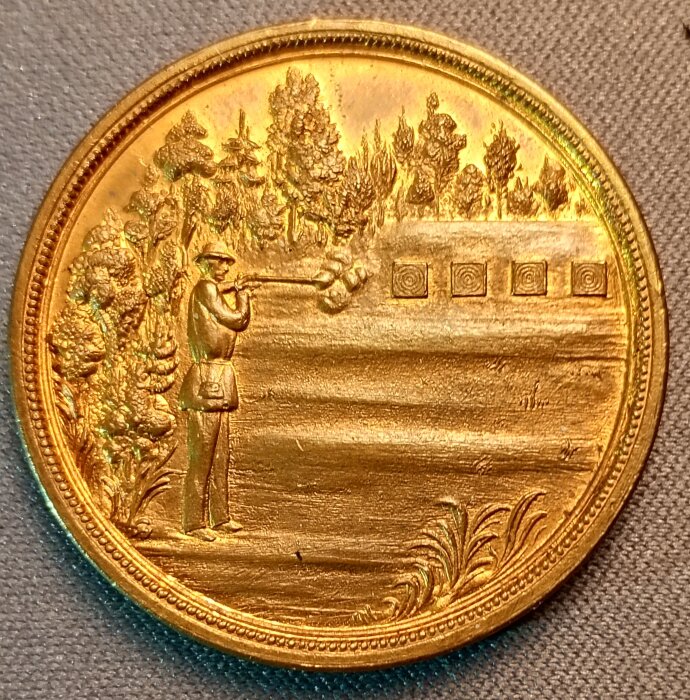 Skyttemärke i guld med motiv av en skytt som siktar mot mål i en skogsmiljö, omgiven av detaljerade träd.