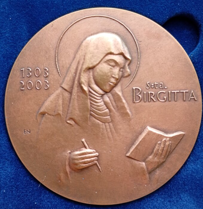Medalj av heliga Birgitta, visar en kvinna i slöja med ett skrivblock och penna, texten "S:ta Birgitta" och årtalen 1303-2003.