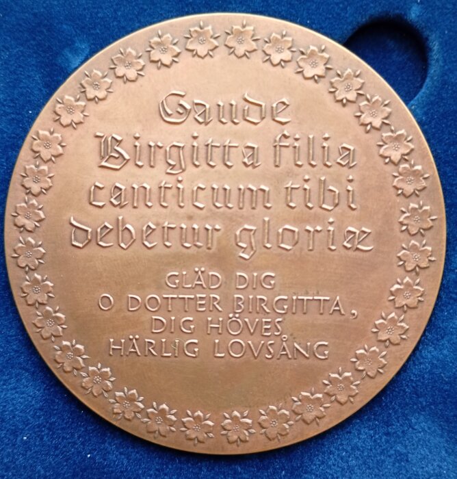 Medalj av heliga Birgitta med blomdekoration och latinsk text, placerad på blå bakgrund.