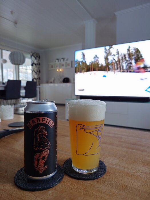 En ölburk och ett glas med skummande öl står på ett bord i ett vardagsrum med en påslagen TV i bakgrunden.
