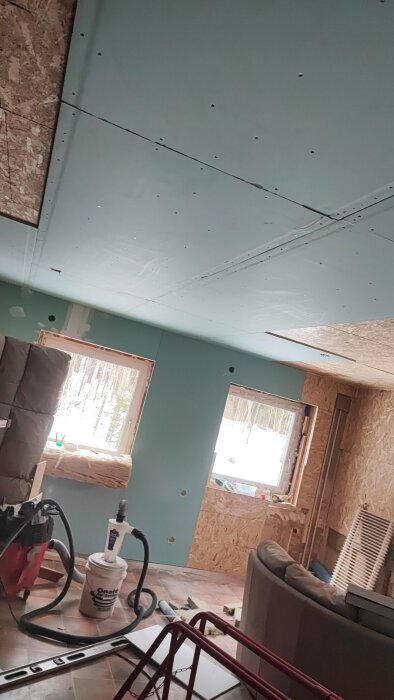 Rum under renovering med gipsplattor på taket, verktyg på golvet, isolering vid fönstret och ett soffhörn synligt.
