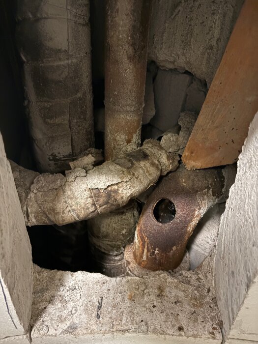 Rostiga, isolerade rör och en betongöppning med synlig skada i en vägg.