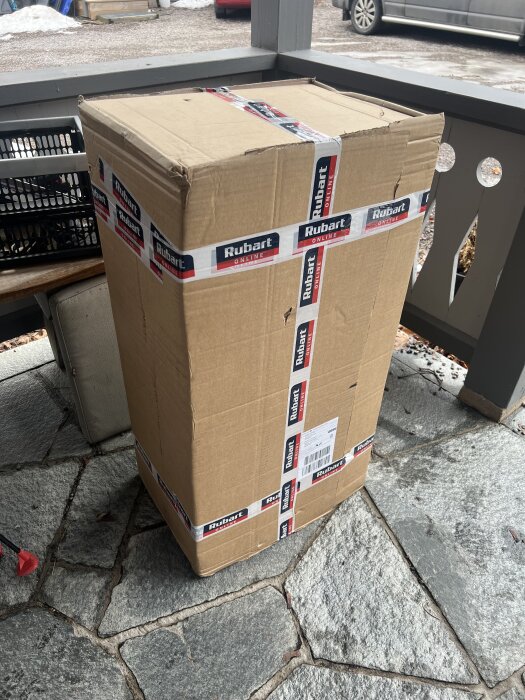Stor kartong märkt med "Rubart Online" står på en stenterass, omgiven av andra föremål och vinterlandskap i bakgrunden.