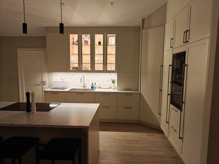 Modernt kök med ljusa skåp, marmorbänkskiva och inbyggd ugn. Skåp med vitrindörrar och belysning visar glas och prydnader.