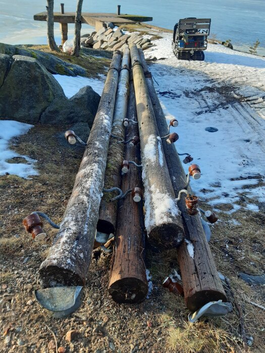 Gamla telefonstolpar ligger staplade på marken nära en sjö, omgivna av snö och stenar, med en skoter i bakgrunden.