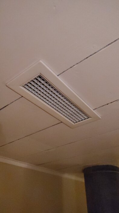 Ventilationsutblås i tak med gallermönster bredvid en mörk rökgång från vedkamin, del av ett värmeåtervinningssystem.