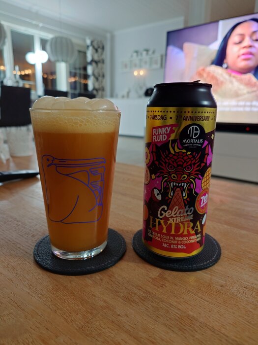 Glas med orange suröl och burk med etikett från Funky Fluid, placerade på ett bord i ett vardagsrum med TV i bakgrunden.