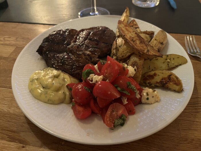 Grillad biff med klyftpotatis, körsbärstomatsallad och bearnaisesås på en vit tallrik.
