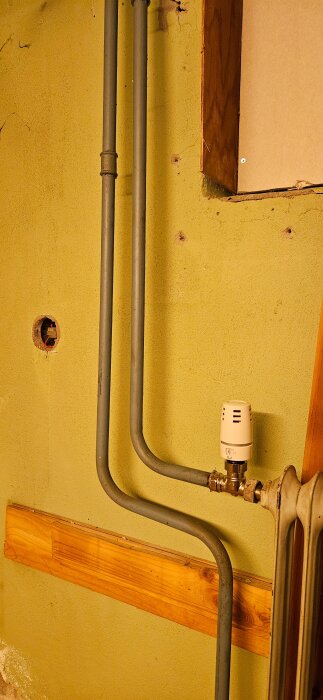 Gammalt element med metallrör i källare, kopplade till en vit termostat på grön vägg med träpaneler och synligt röruttag.