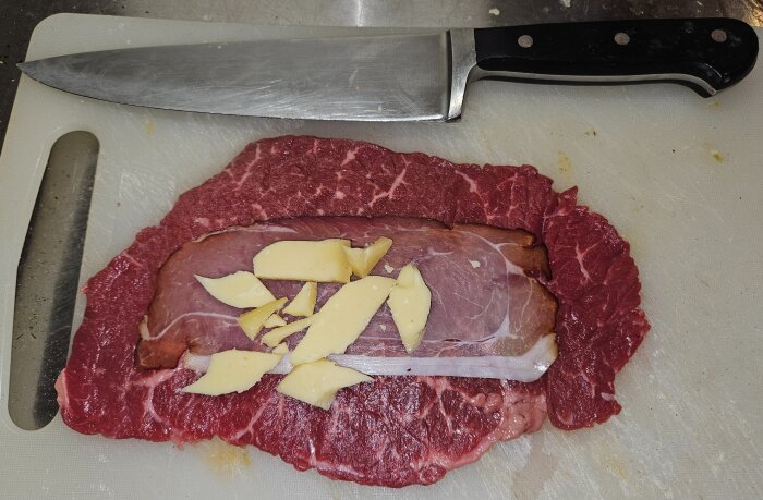 Cordon Bleu-förberedelse med lövbiff, Schwarzwald-skinka och Gryere-ost på en skärbräda, bredvid en kniv.
