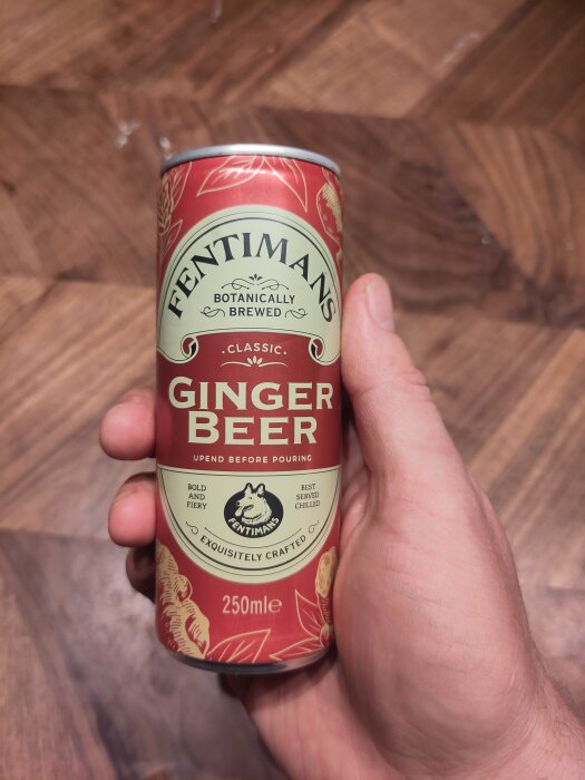 En hand håller en burk av Fentimans klassiska ingefärsöl på ett trägolv.
