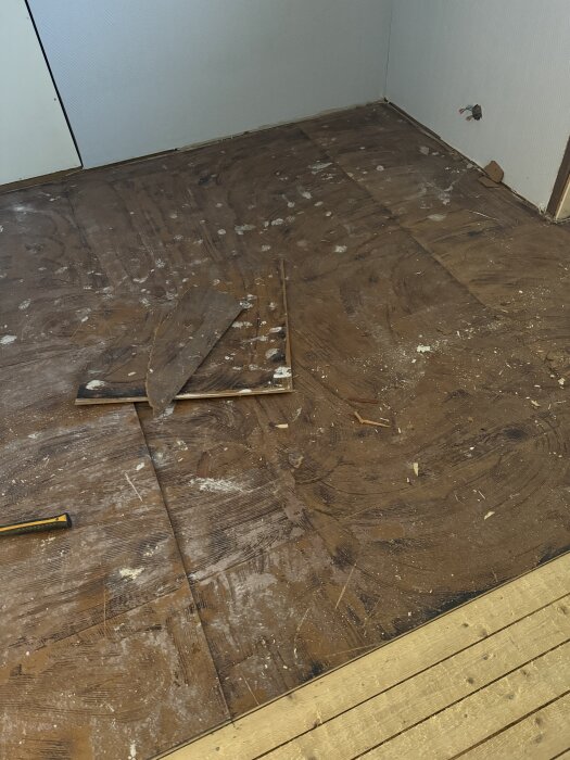 Trägolv under renovering med gamla plankor, sågspån och verktyg. Det finns en väggkontakt utan skydd tätt intill hörnet.
