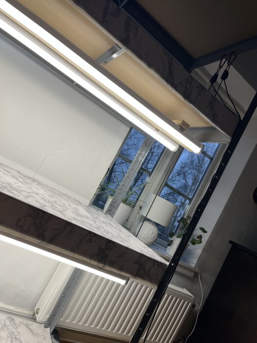 Hyllplan med vaxduk och lysrör i lagerhylla, fönster med växter i bakgrunden och karbinhakar för upphängning synliga ovanför.