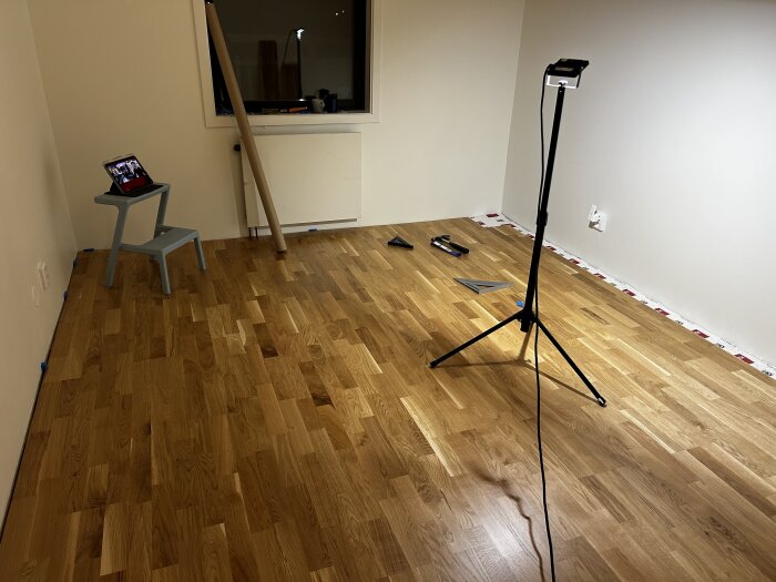 Rum med nylagt trägolv, en stege med surfplatta, stående lampa och verktyg på golvet. Fönster och vit vägg synliga.