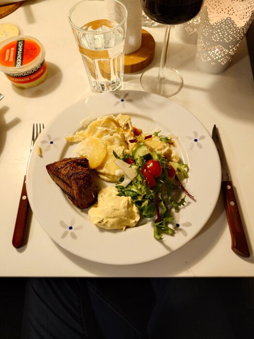 Grillad rostas och potatisgratäng på en tallrik med sallad och sås, serverad med ett glas vatten och vin.