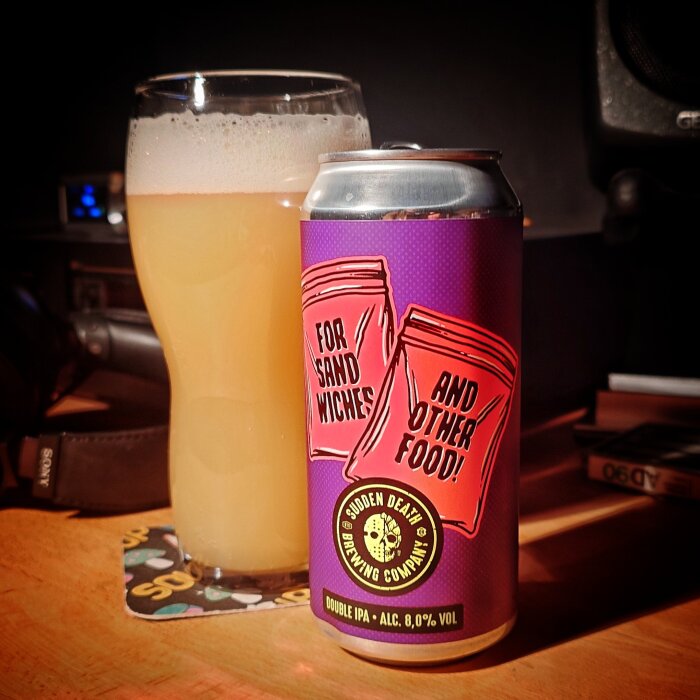 Ölburk med etikett "For sandwiches and other food" från Sudden Death Brewing Company, bredvid glas med ljus öl på bord i svagt ljus.