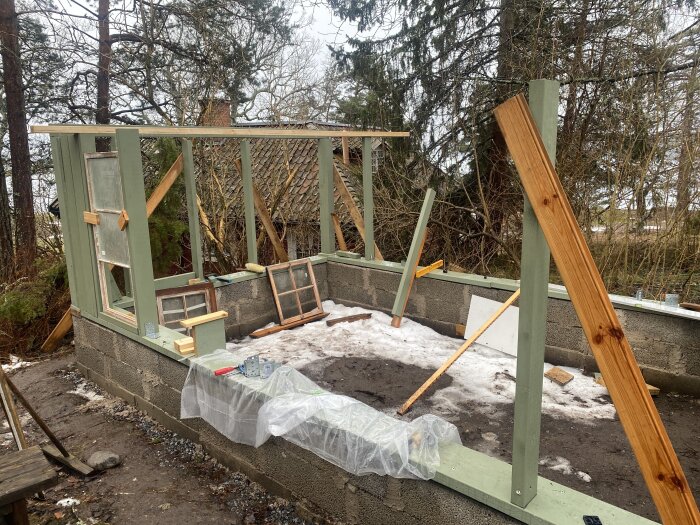 Byggnation av växthus med betongmur och trästolpar, några fönster lutade mot muren och plast på marken.