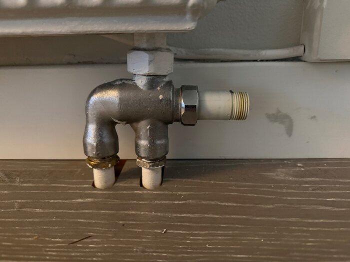 Metallrör och kopplingar under en radiator fästa vid en vägg, med synliga gängade anslutningar och vitmålad bakgrund.