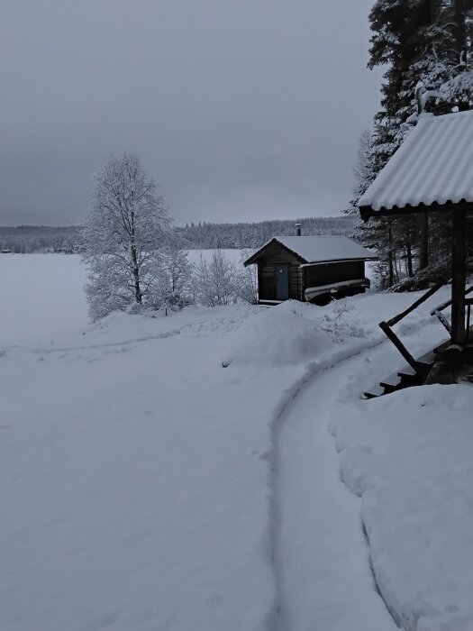 Snötäckt landskap med stuga och staket vid sjö, omgiven av snötäckta träd och en stig i snön. Grå himmel i bakgrunden.