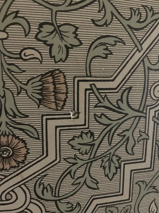 Närbild av en tapet med ett detaljerat blommönster i grönt och brunt, samt en liten skada eller reva i ytan.