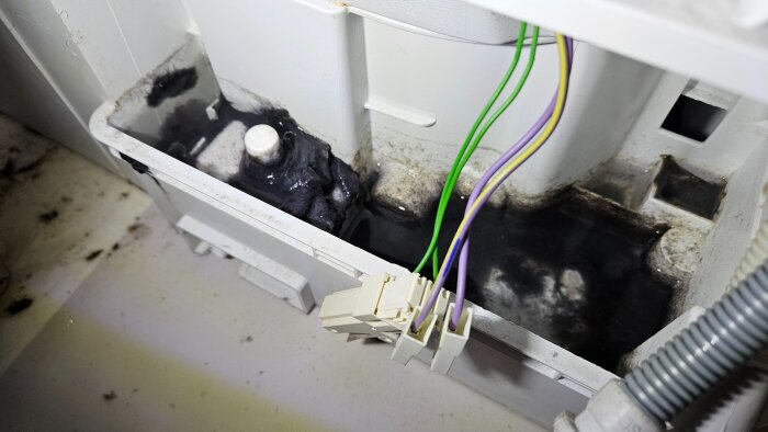 Bild på baksidan av en torktumlare med dammansamling och vattenläckage runt elektronik och kondensator, vilket orsakar överhettningsproblem.