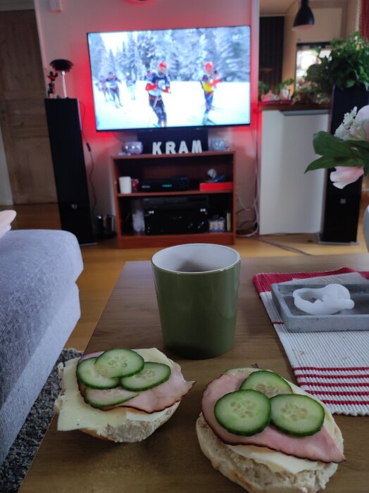 Mackor med skinka och gurka på ett bord framför en TV som visar skidcupen; grönt kaffekopp bredvid och inredning i vardagsrumsmiljö.