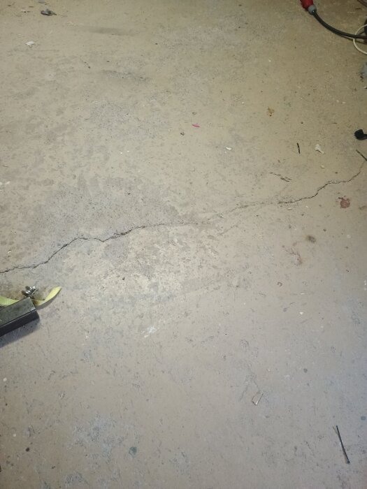 Sprucket och fläckigt gammalt betonggolv i en verkstad, med diverse föremål längs kanterna och en tydlig lång spricka i mitten.