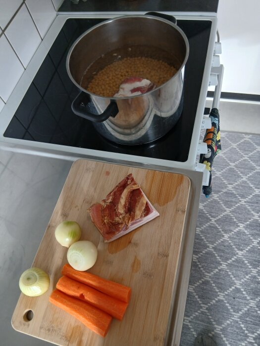 Gryta med ärtor och kött på spisen, bredvid en skärbräda med lök, morötter och fläskbitar.