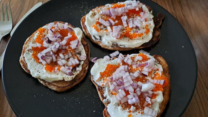 Surdegspannkakor toppade med gräddfil, rödlök och röd stenbitsrom på en svart tallrik.