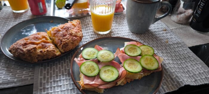Frukost med skinksmörgås med gurkskivor, ostfralla, ett glas apelsinjuice och en kopp te på ett bord med underlägg.
