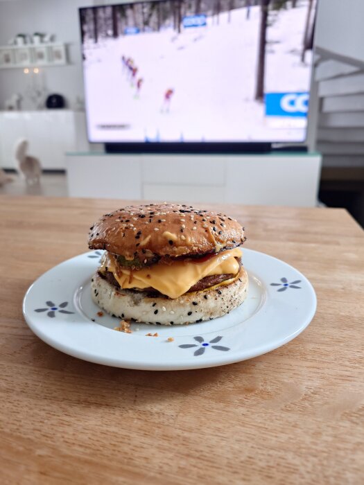 Hamburgare med sesamfrön och smÀlt ost pÄ tallrik framför TV med skidÄkning.