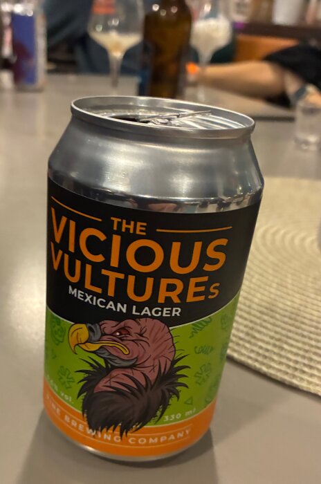 Ölmärket "The Vicious Vultures" Mexican Lager på en burk, fotograferad på ett bord med diverse drycker i bakgrunden.