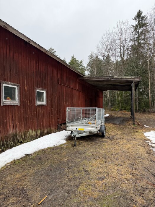 Röd äldre lada med flagnande färg, släpvagn framför, och en del snö kvar på marken. Skog i bakgrunden.