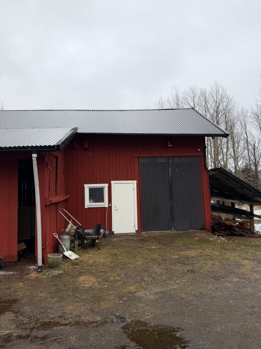 Röd lada med grått tak och vita dörrar, omgiven av jord och lite snö. Några träd skymtas i bakgrunden under en mulen himmel.