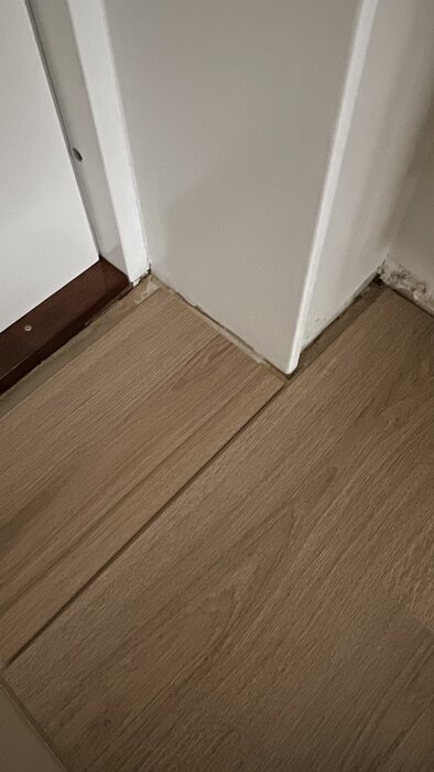 Hörn av rum med laminatgolv och en träbräda vid väggen med en smal springa längs kanten. Diskuteras om kapning eller list är bäst.